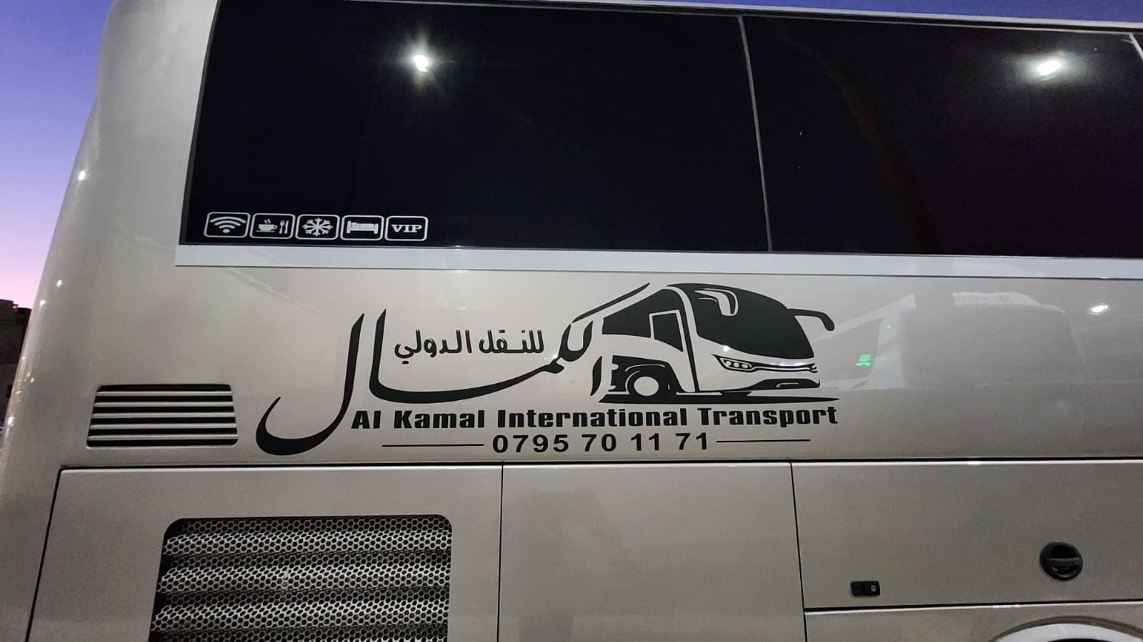 جانب حافلة بيضاء لشركة الكمال للنقل الدولي مع شعار وأيقونات خدمات تحت سماء الغسق.